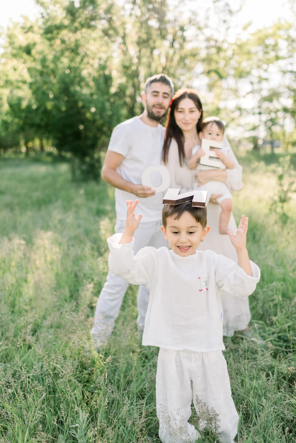 Un băiețel ridică mâinile în joacă, cu familia în spatele lui, într-o grădină verde.